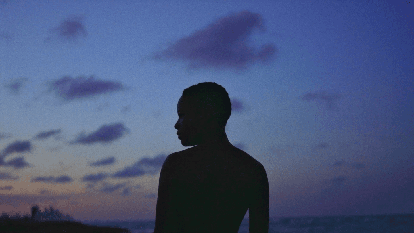 A Black person stands silhouetted against an early evening sky - the sun setting in the far distance. They look to their left.
