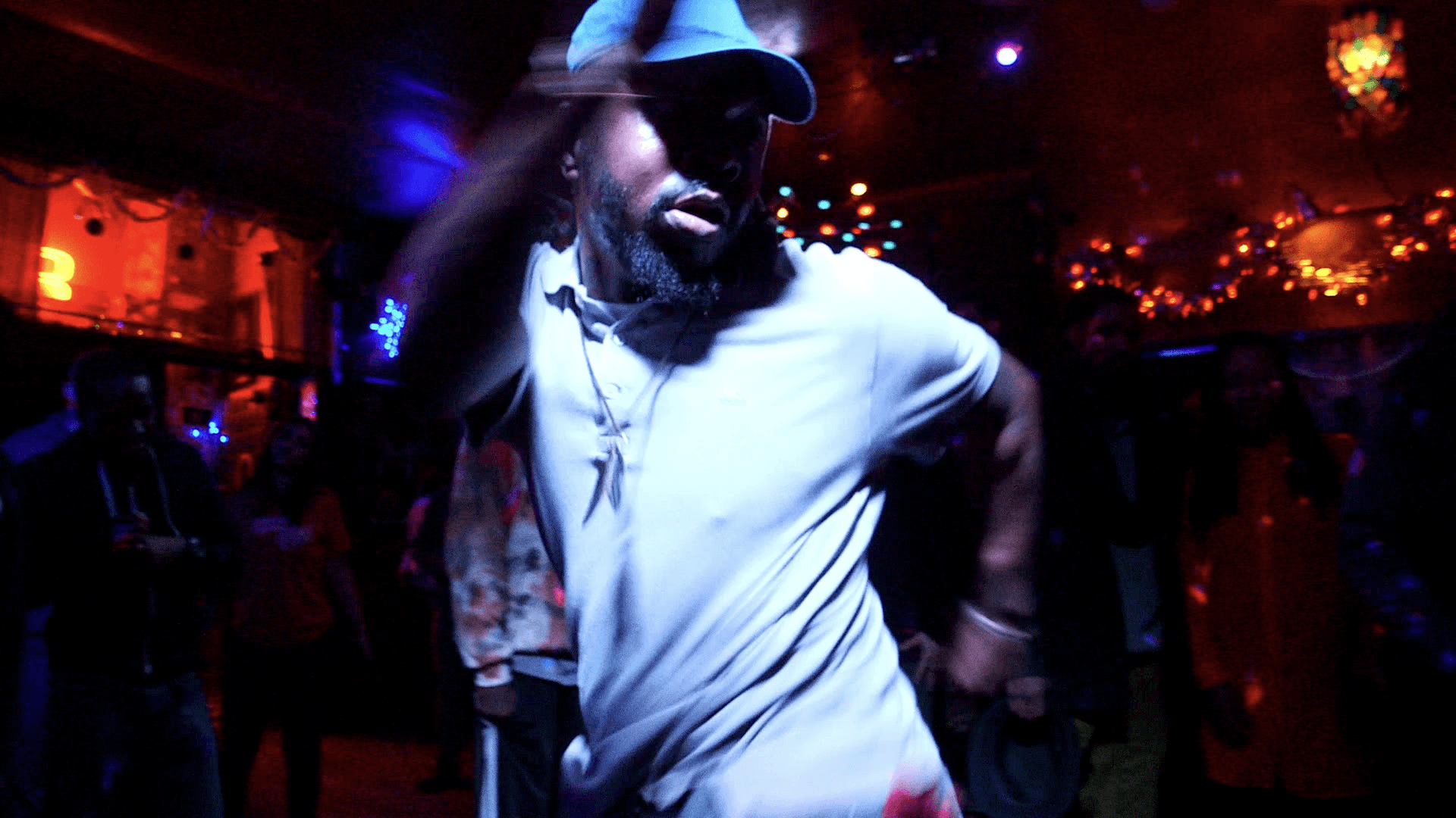 A photo of a Black person dancing at a club. They are wearing a white polo and a baseball cap. The photo is slightly blurry, there are colorful string lights glowing in the background.