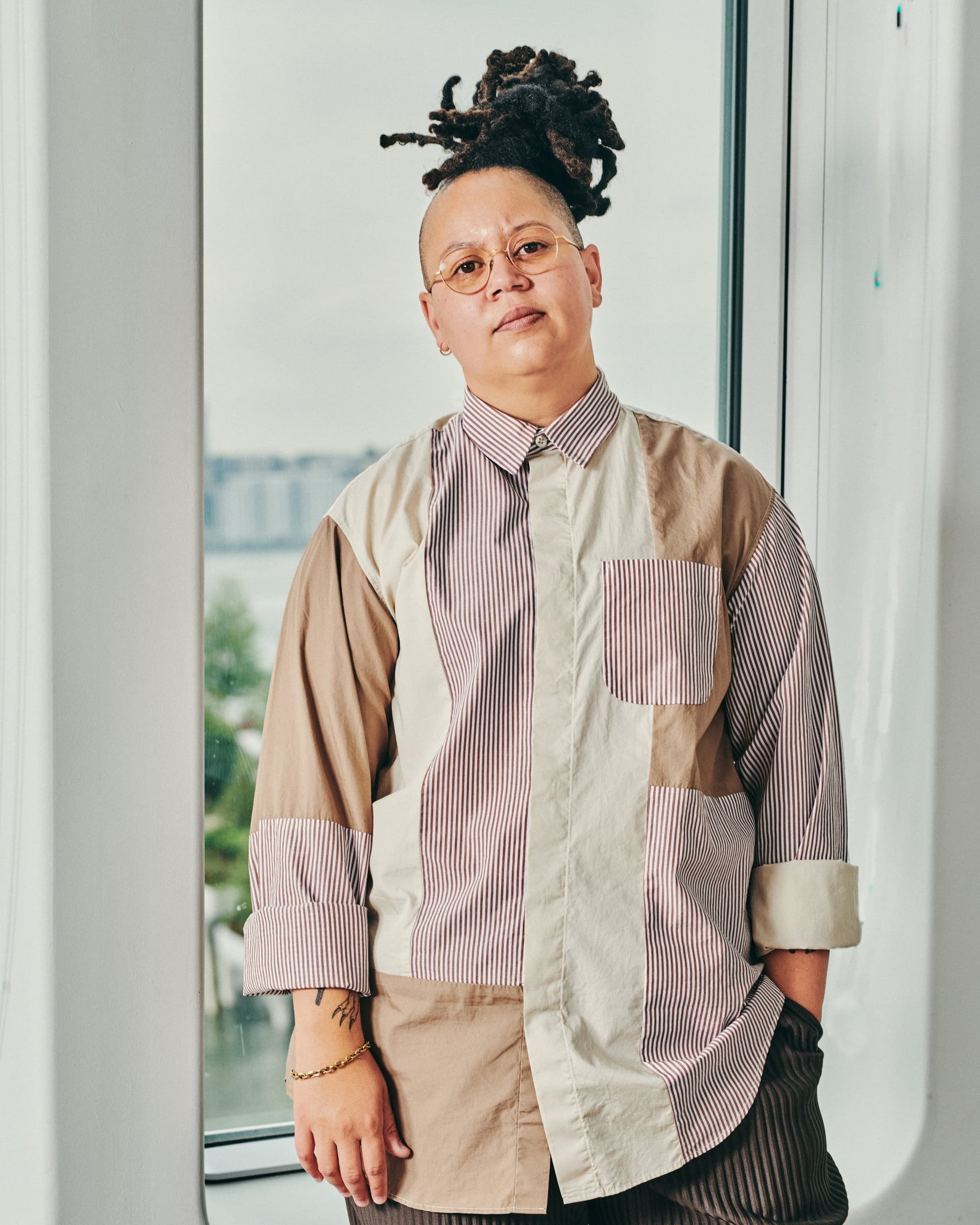 A headshot photo of Meg Onli. They have a light skin-tone. The sides of their head are shaven and they have long locs tied up into a bun. They are wearing a mutli-patterned button down shirt.