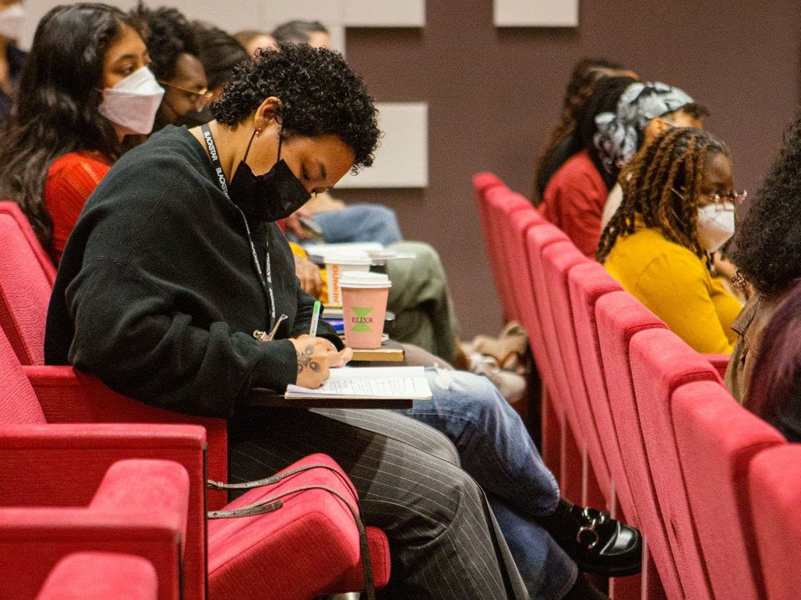 A photo of group of people in auditorium-like setting. The person is the foreground is taking notes.