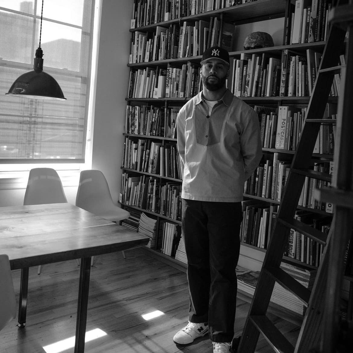 A photograph shows a black and white portrait of Sean-Josahi Brown standing in a sunlit room with a table, chairs, and a wall of book shelves behind him.