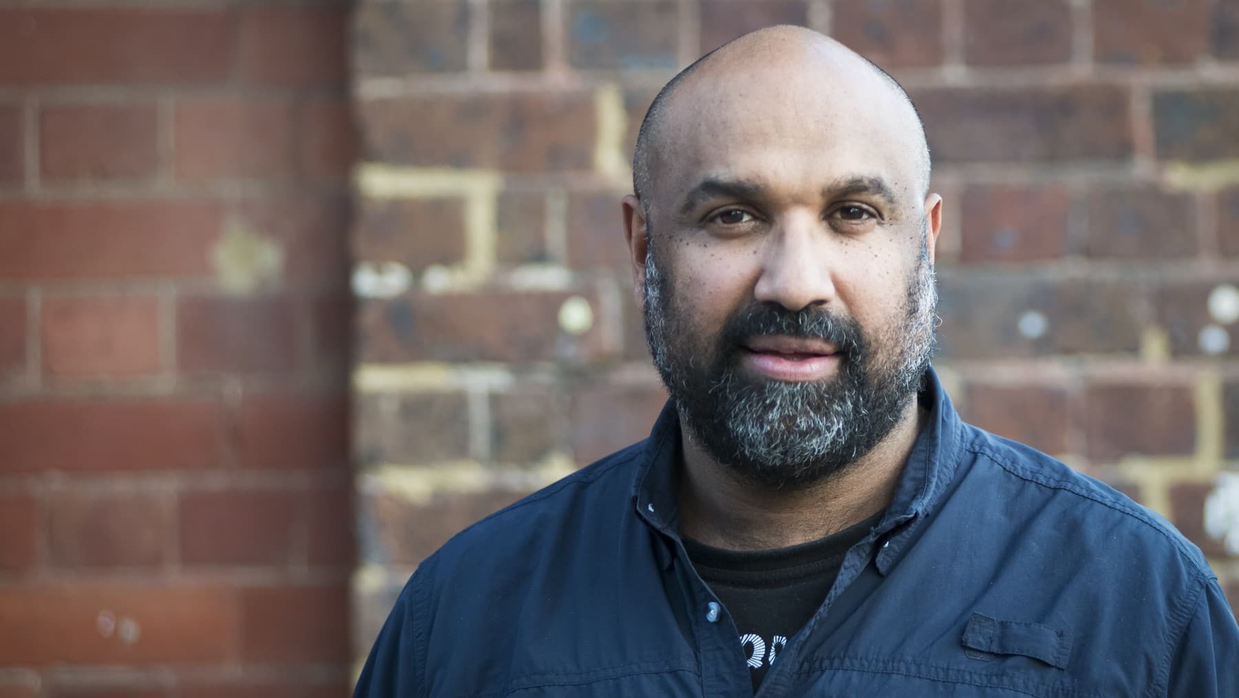 A picture of a man named John Harvey against a brick wall. He is bald with a short, graying beard and he is wearing a dark navy-blue jacket over a black shirt with white text, partially obscured. His expression is calm and slightly serious, with his eyes looking directly at the camera.