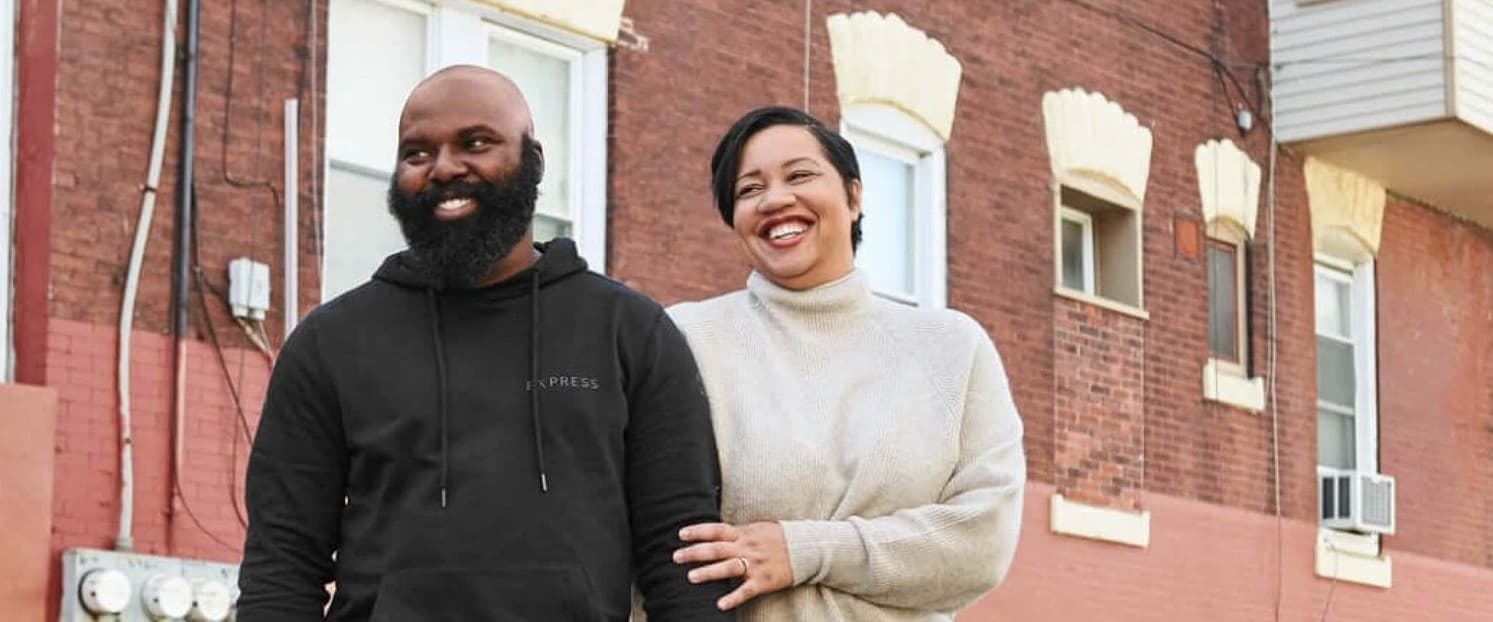 A photo of Omar Tate and Cybille St.Aude Tate. Omar is a Black man, he is bald with a large beard. He is wearing a black sweatshirt and blue jeans. Cybille is a Haitian woman, she has short black hair. She is wearing a white knit sweater and black pants. Omar and Cybille are holding hands and smiling.