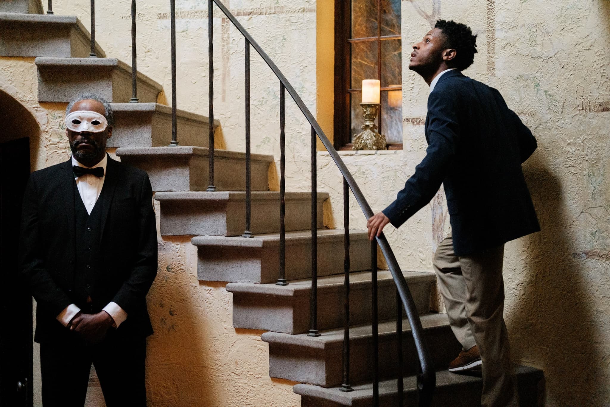 A still from The Freedom to Fall Apart shows two Black men in a mansion-like setting. They are dressed in suits. One is wearing a white mask. The other, who is not wearing one, is looking up a staircase with a curious expression.