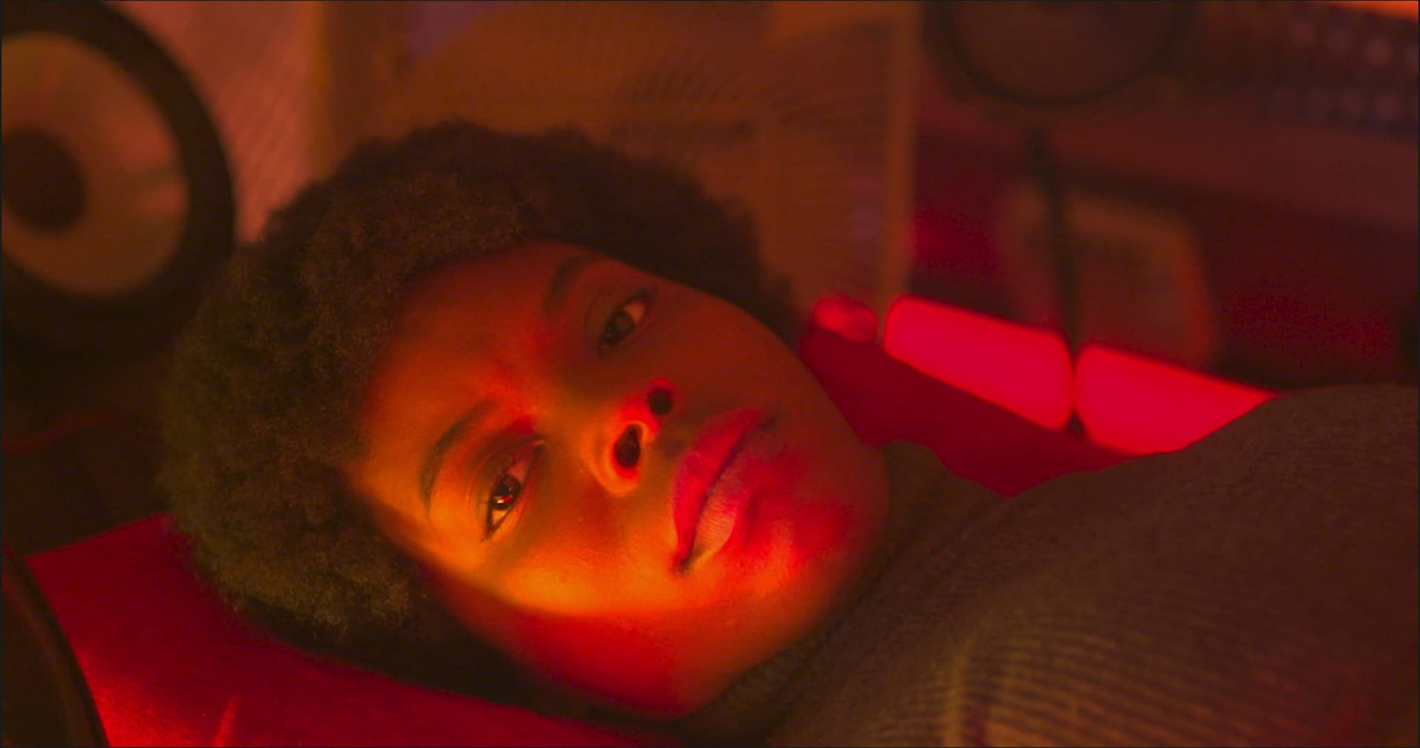 A still from The Love Machine shows a young Black girl laying down. There is a soft red glow on her face.