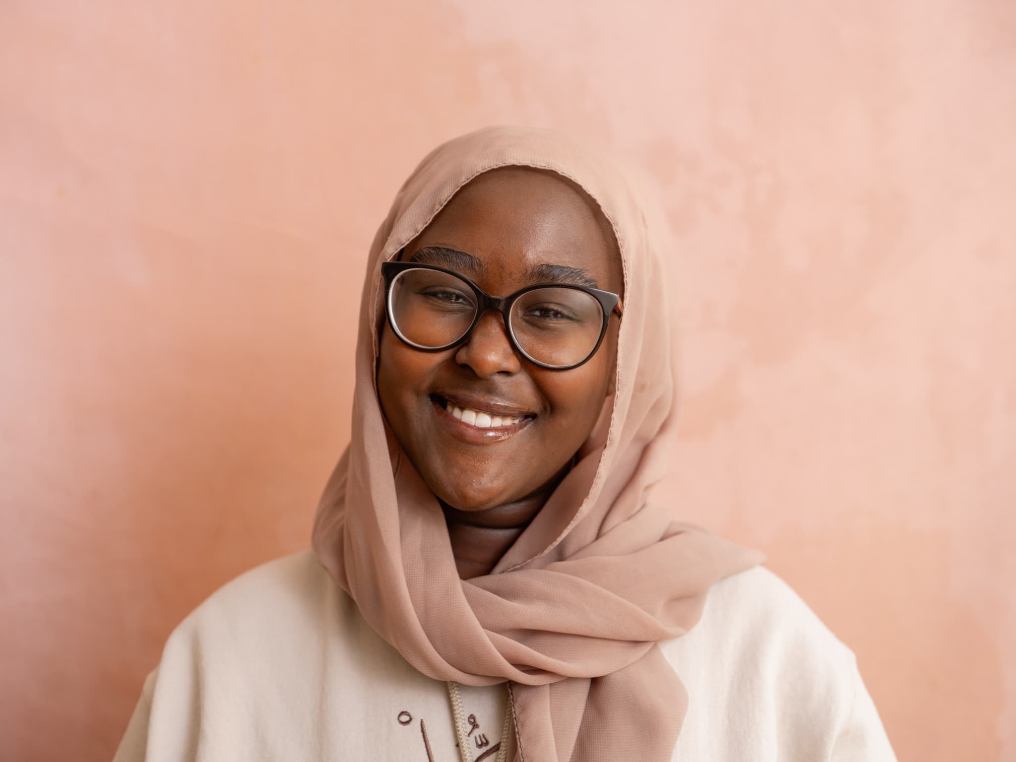 A headshot of Amina. She is a young Black woman. She is wearing a hijab and glasses. She is smiling at the camera.