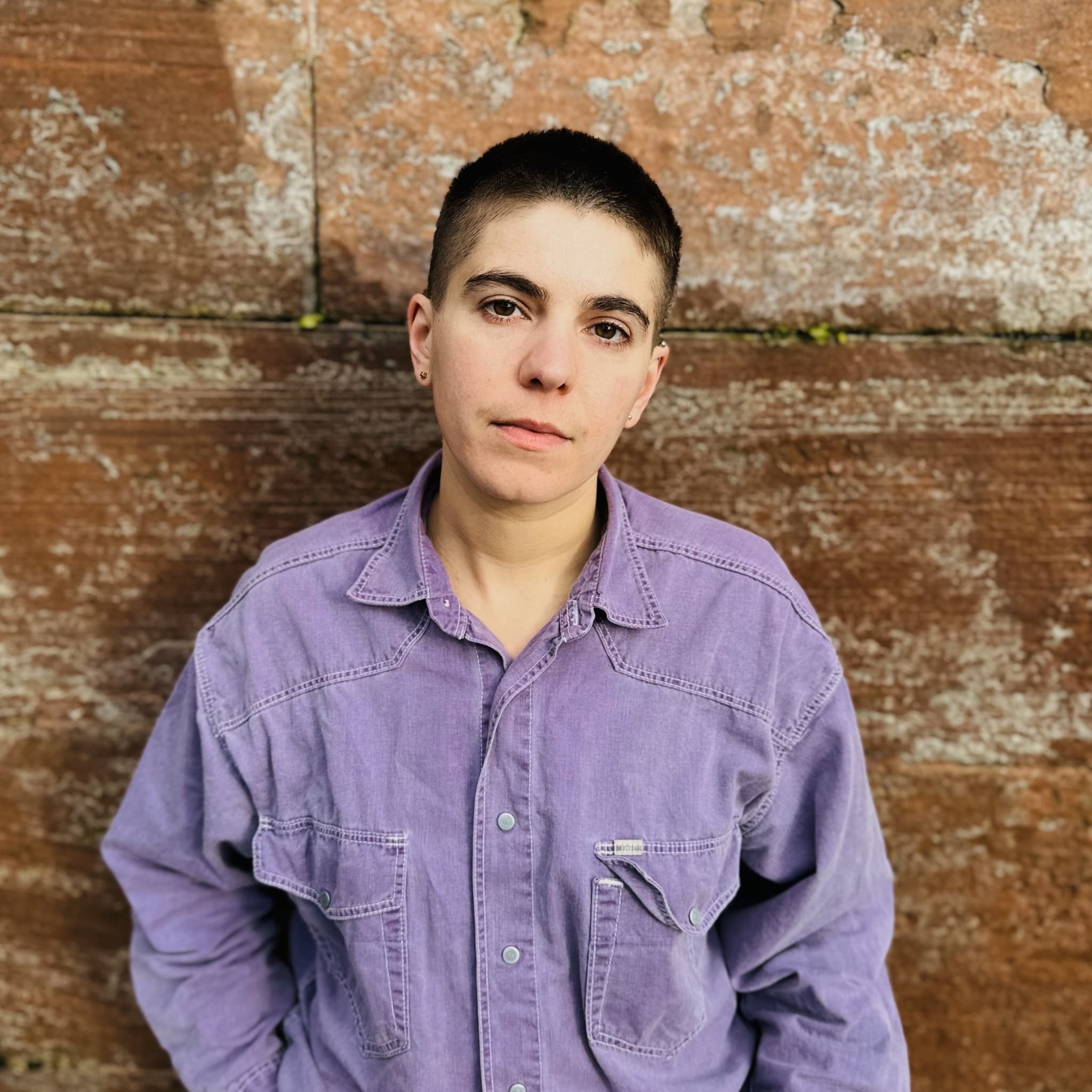 A picture of a person named Emilia Beatriz. They are wearing a deep lilac colored jean jacket and blue denim jeans, against a worn rusty background. They have a brunette buzzed haircut and are looking at the camera fondly with their head tilted slightly.