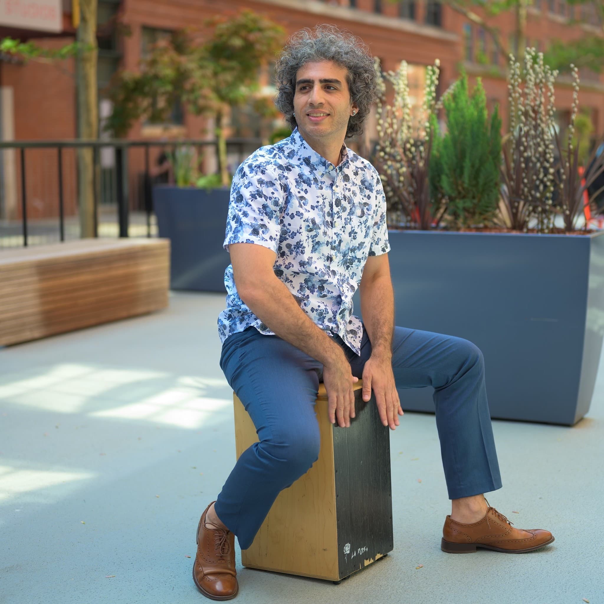 A picture of a man named Tareq Rantisi. He has graying curly hair, is wearing brown slacks, and is sitting in a straddle position atop a wooden block on an outdoor patio in the daytime. He wears a blue and white floral short sleeve button down top with matching solid blue pants. Looking off to the side in curiosity he has his hands resting on the block and his feet pointed outwards.