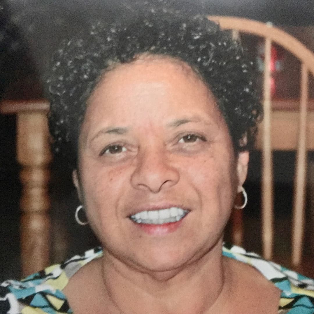 A picture of a person named Ada Gay Griffin. She is smiling and faced directly towards the camera. She has brown short curly hair and two small hoops on her ears. She wears a blue and green geometrically shaped top against an indoor residential background.