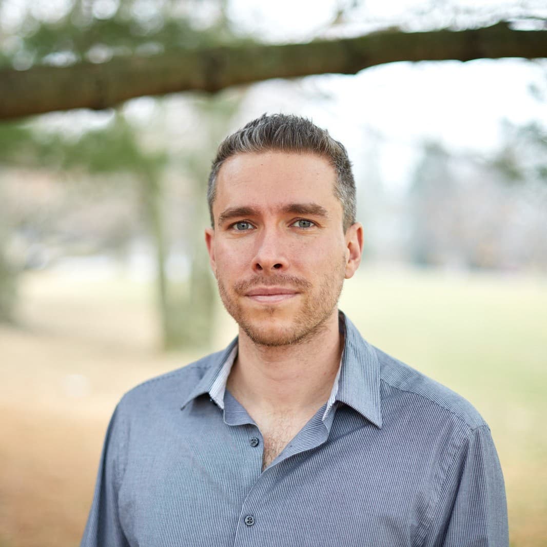 A picture of a man named Daniel Glick. He has blue eyes matching the blue button down top he wears with an open collar. He has short graying hair and a faint beard and mustache. With minimal expression he stares into the camera against an outdoor background.