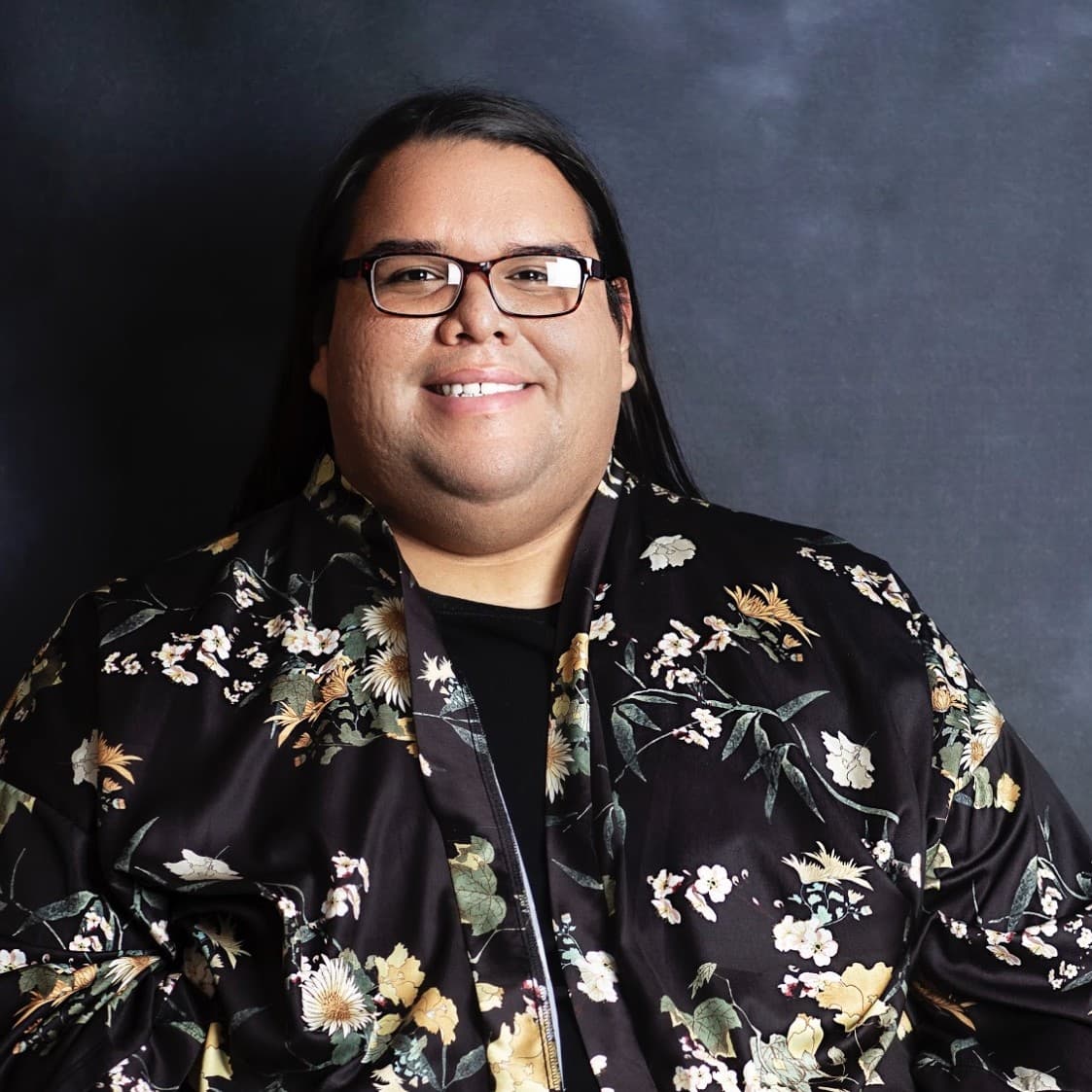 A picture of a man named Ivan Mcdonald. He is sitting down and has long black silky hair pulled back, and is wearing black satin floral top. He wears black glasses and is smiling happily towards the camera against a dark gray background.