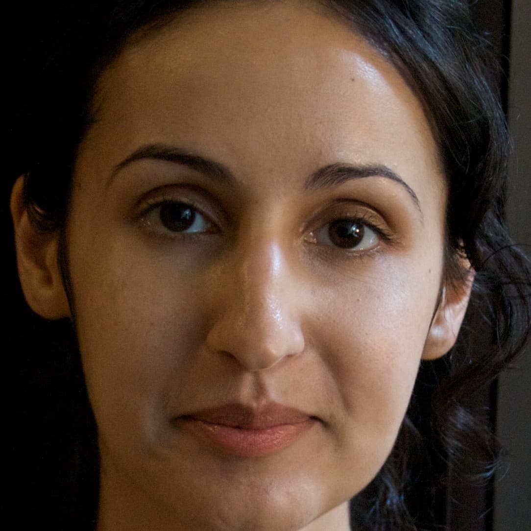A picture of a woman named Mariam Ghani from the neck up focused intently on the camera. Her dark wavy hair is pulled back, showcasing her blank expression and downturned faint smile.