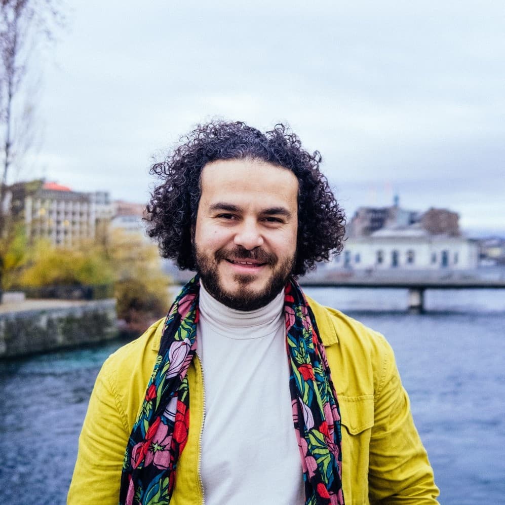 A picture of a man named Mohamed Jabaly outside next to a lake with buildings and a bridge in the background. He stands out from the blue washed scenery in his bright yellow open sweater and colorful floral scarf. He has short brown curls, facial hair, a white turtleneck, and a happy grin towards the camera.