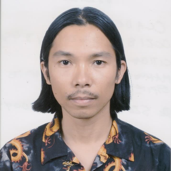 A picture of a man named Miko Revereza. He is staring straight towards the camera, his body completely centered. He has straight sleek short black hair, cut in a straight bob and tucked behind his ears. He is wearing a black button down top with fiery orange dragons, with a light mustache just above his soft faint smile.