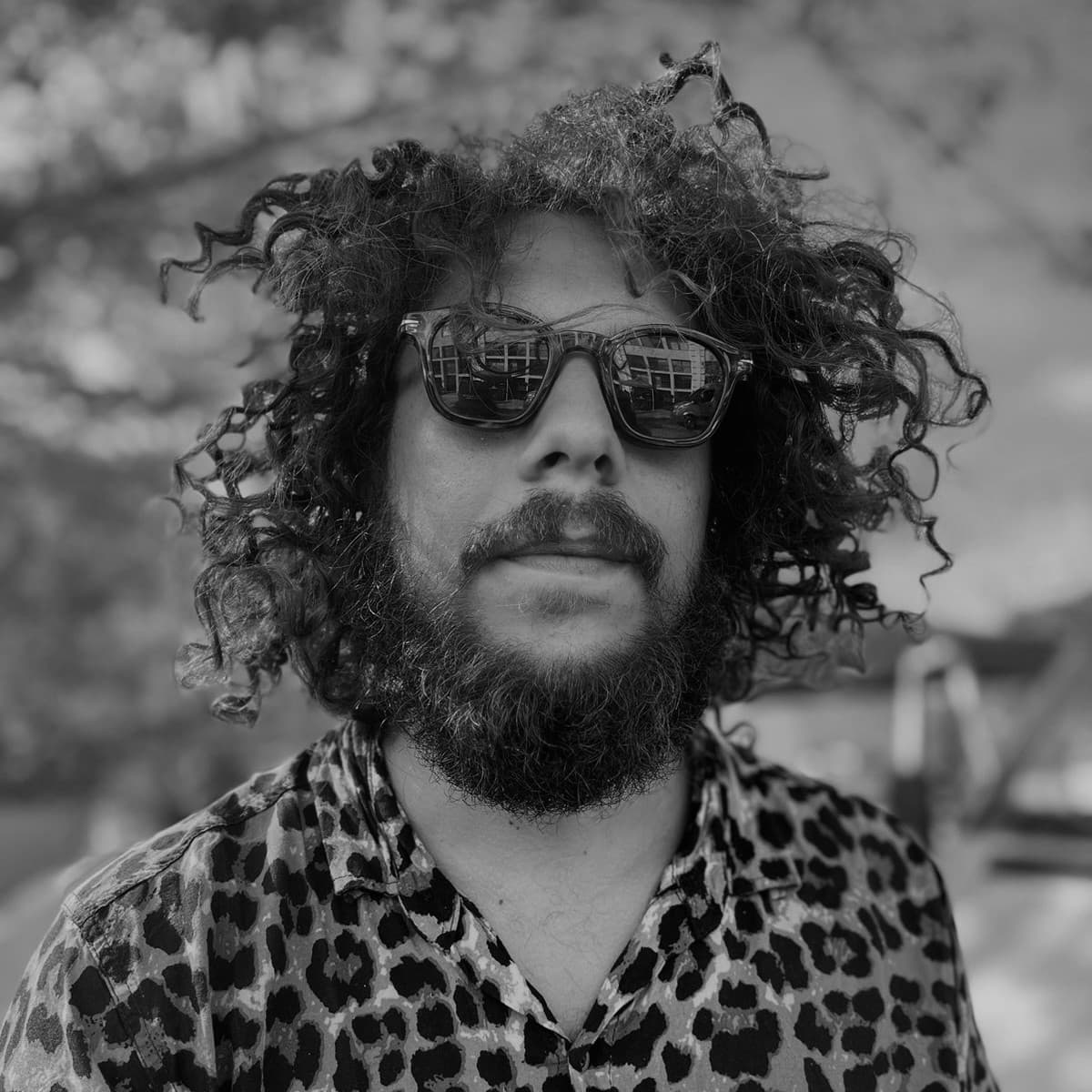 A black-and-white picture of a man named Lucas Rossi Dos Santos. He has a thick dark mustache, beard, and curly hair, with several stands spiking in different directions. He wears a cheetah print button down top, dark sunglasses, and pursed faint smile against an outdoor background.
