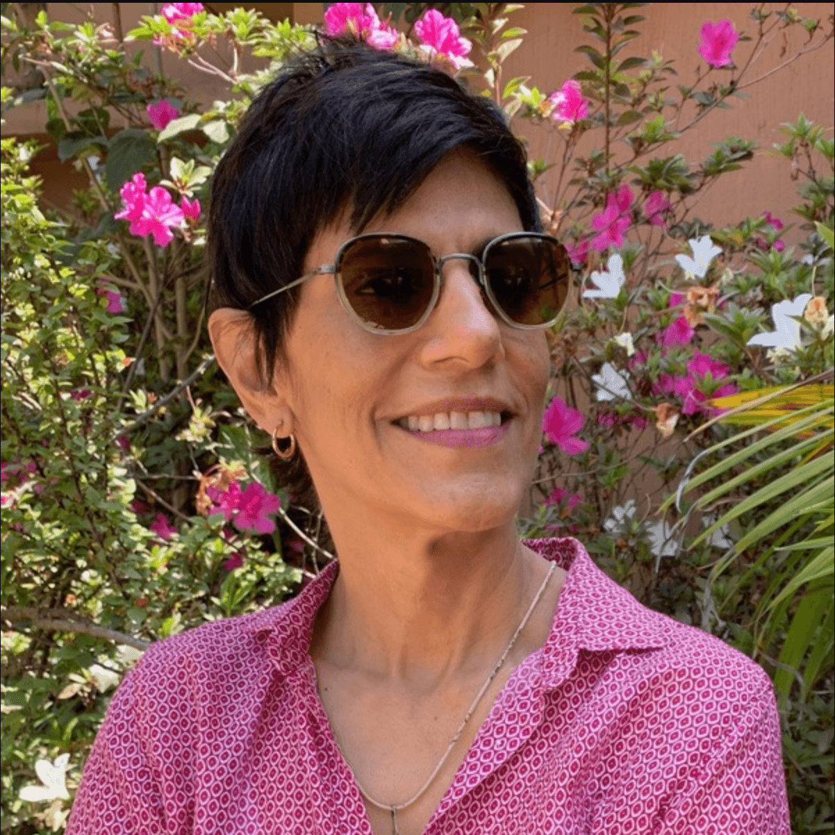 A picture of a woman named Meena Nanji posed gracefully surrounded by a garden of greenery and pink florals, her neck adorned with a necklace. She wears a matching pink button down top, brown sunglasses matching the color of her short hair and smiles warmly turned looking off towards something to the right.