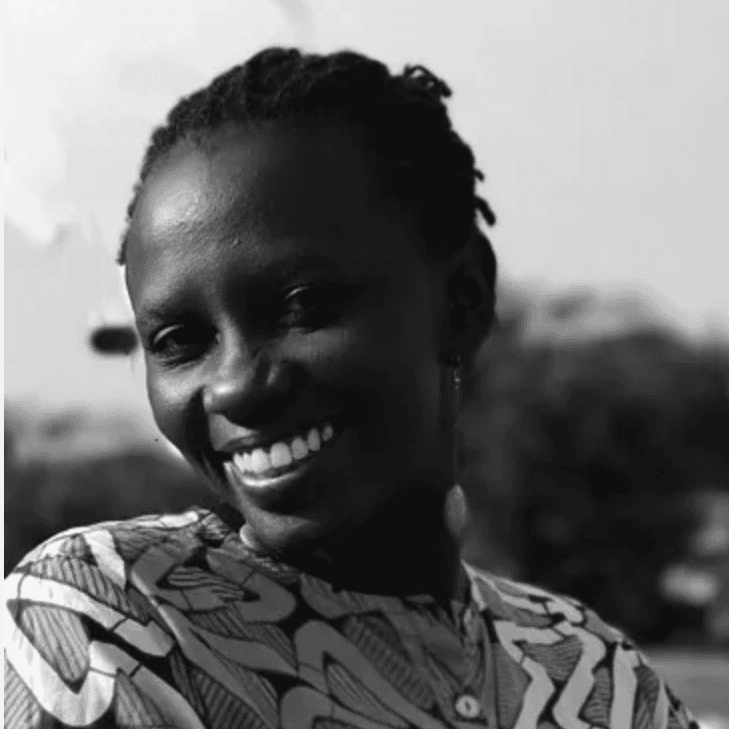 A black-and-white picture of a woman named Zippy Kimbundu. She has a bright wide smile in contrast to her dark skin, wearing an intricate shape patterned button down top. She has tightly coiled 4c hair pulled back against an outdoor background.