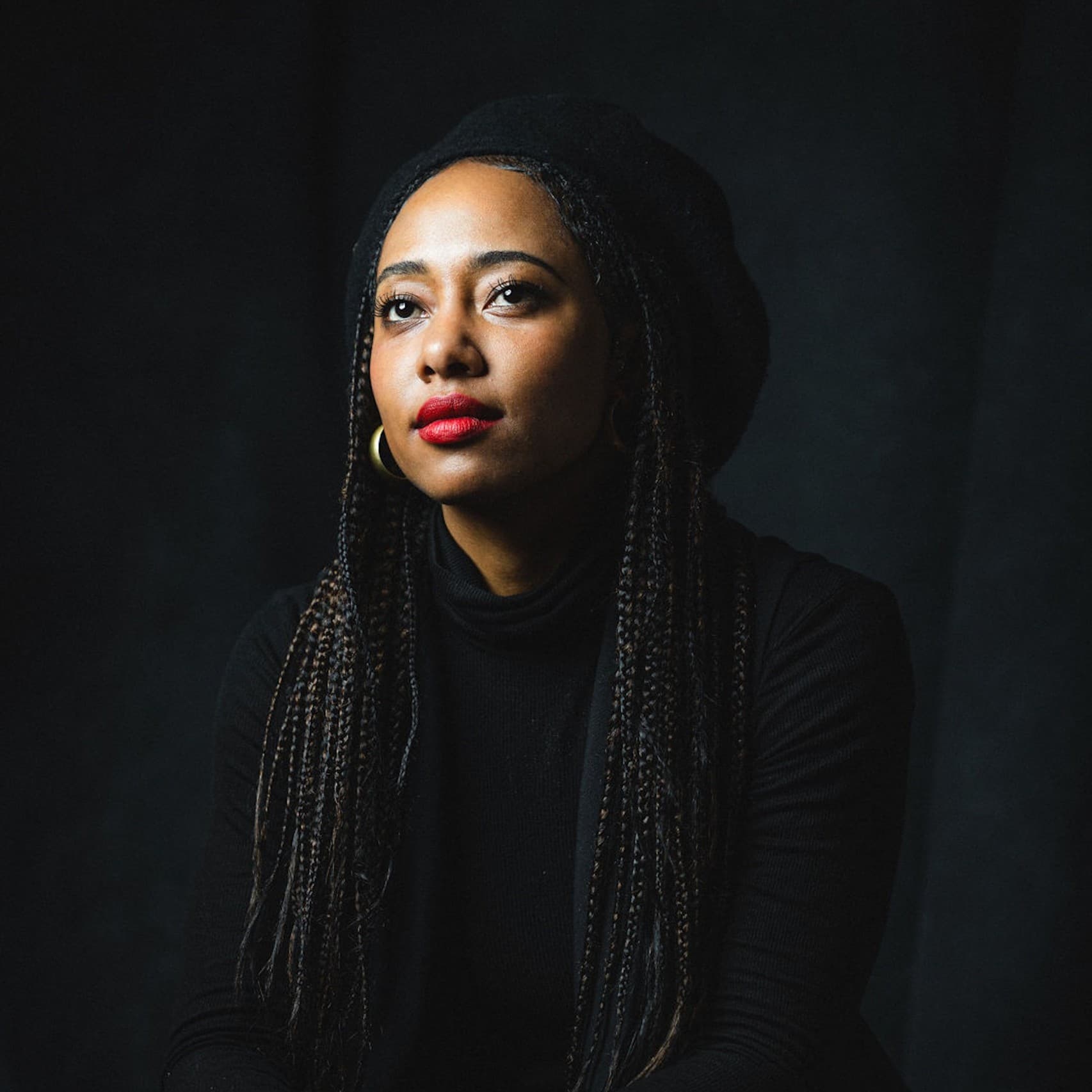 A picture of a woman named Contessa Gayles seated against a black backdrop. She wears a fully black outfit consisting of a beret and turtleneck, barely visible against the dark background. Her long, small, braided hair falls over her shoulders as she holds a calm, contemplative expression gazing slightly upwards and to the side. Her bold red lipstick stands out prominently against her dark attire, and her hands are clasped loosely in her lap.