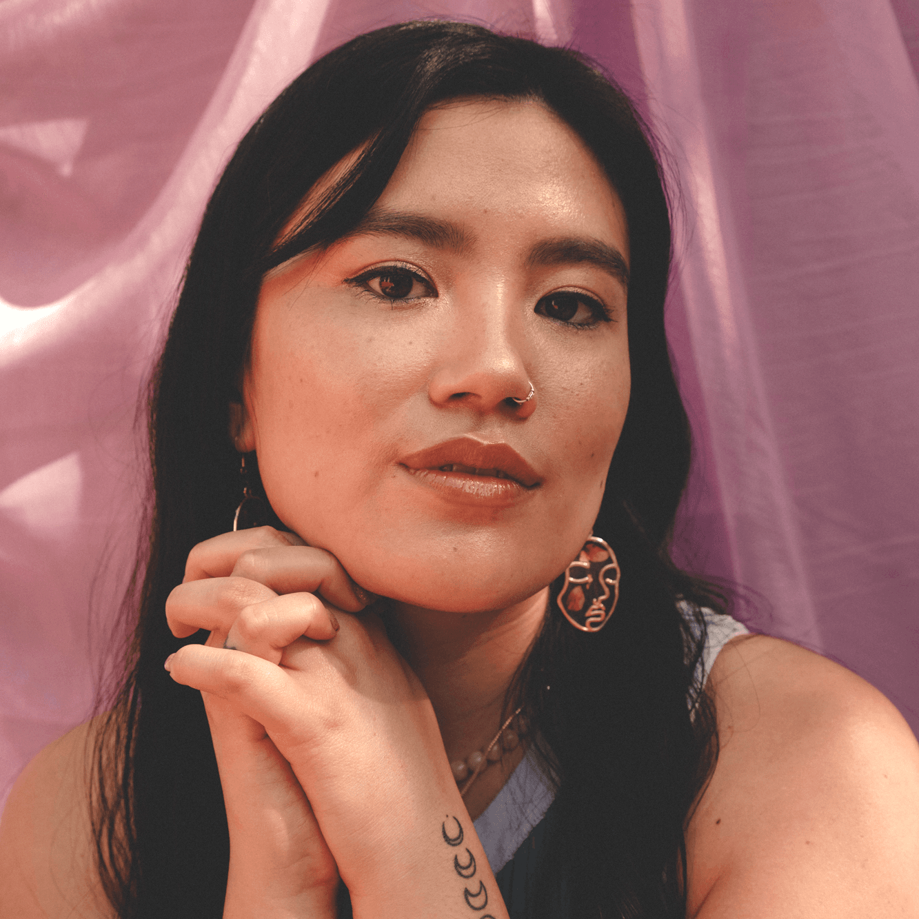 A picture of Jalena Keane-Lee in a serene and contemplative pose against a backdrop of soft, flowing purple fabric. Her long, black hair frames her face, and she is wearing a pair of gold, abstract face-shaped earrings. Her expression is calm and thoughtful, with a slight smile on her lips. She has a nose ring, and her hands are gently clasped under her chin, revealing a small tattoo of moon phases on her forearm.