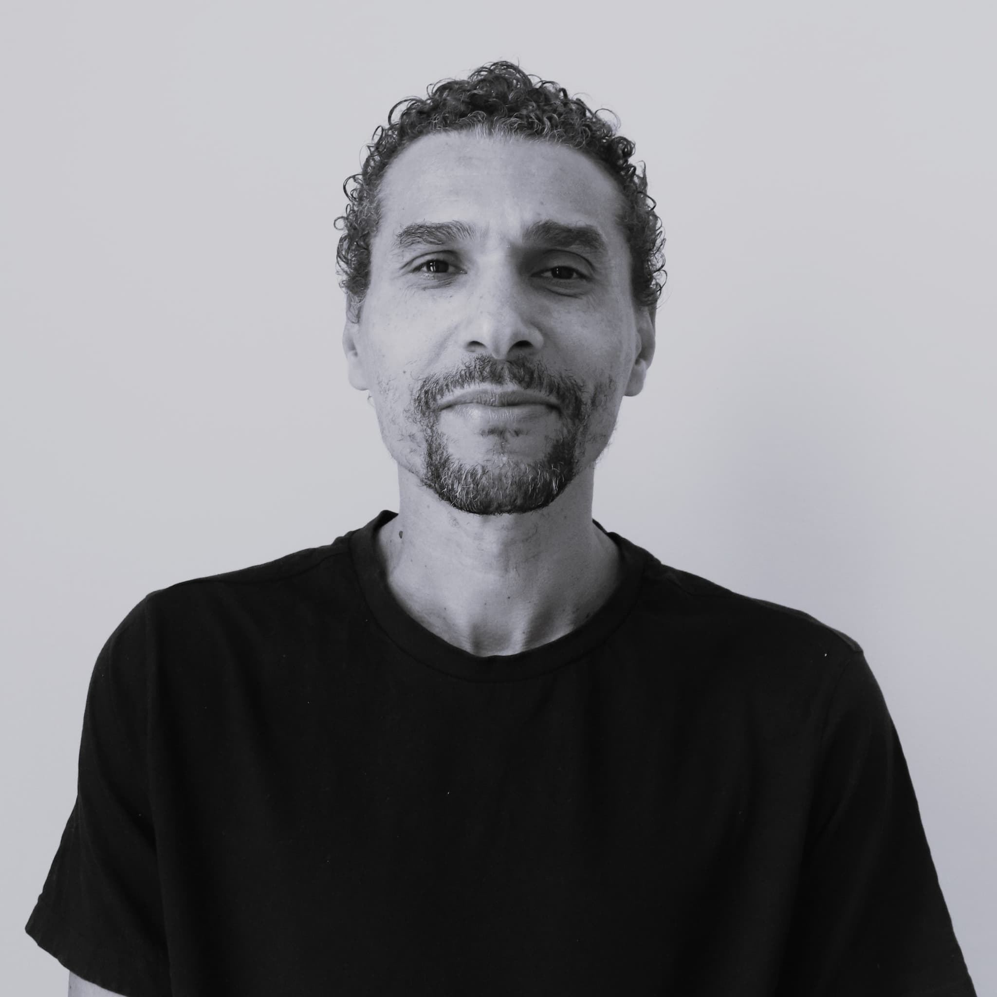 A black-and-white image of a man named Carlos Yuri Ceuninck. He stands against a plain, light-colored background. He has short, tightly curled hair and a neatly trimmed goatee. He is wearing a simple, dark-colored t-shirt with short sleeves. He has a calm and slightly confident expression, with a gentle smile playing on his lips and a relaxed demeanor.