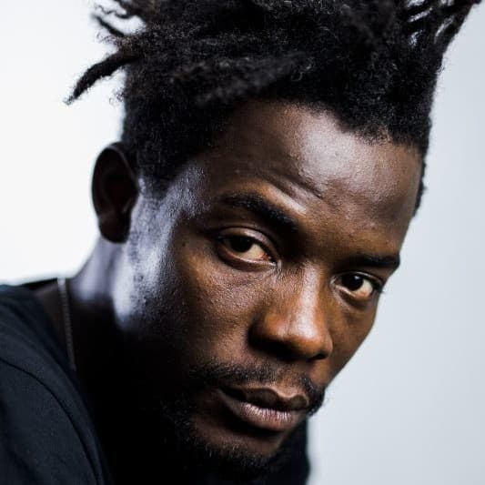 A picture of a man named Pierre Michel Jean against a simple, light gray background. He has a dark complexion, and his hair is styled in multiple short, twisting dreadlocks that are spiked upward and outward. He is wearing a dark-colored shirt, and his facial expression appears serious and contemplative. His eyes are directed towards the camera, with a slight furrowing of the brows and relaxed lips.