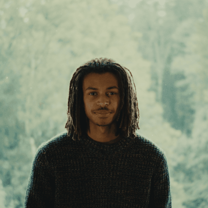 A picture of a young Black man named Damien Hauser, standing in front of a serene blurred rich green background. He has shoulder-length dreadlocks, a neatly trimmed beard, and is wearing a dark, textured woolen sweater. His facial expression is calm, with a small, relaxed smile, and he looks directly at the camera.