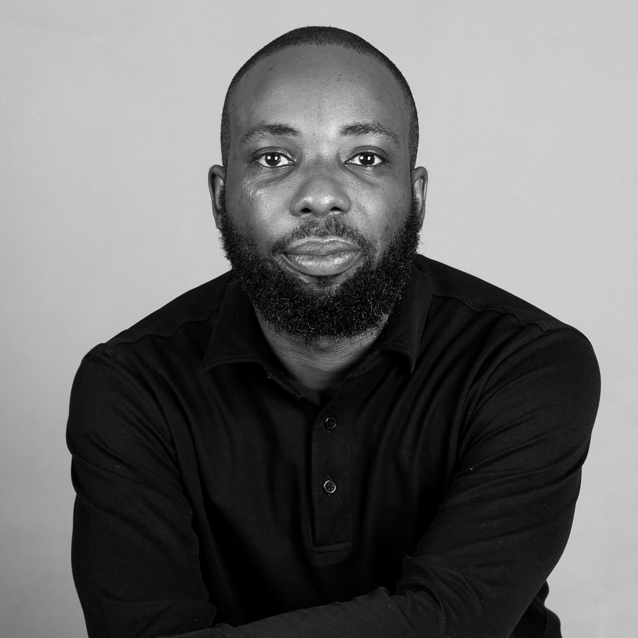 A black-and-white picture of a man named Ishaya Bako. He is centered in the frame against a plain and light-colored background, gazing directly at the camera with a neutral expression. He has a shaved head and a full beard, with closely trimmed sides. He is wearing a dark long-sleeve collared shirt with his arms crossed.