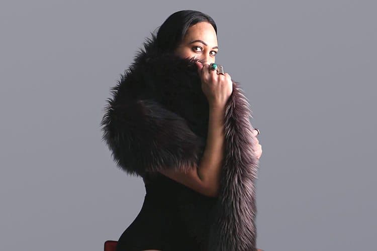 A picture of a transgender person named Ramon Te Wake against a plain gray background. She is dressed in a black, sleeveless bodysuit with a large, plush voluminous fur scarf draped around their shoulders. She holds the fur scarf close with one hand covering her mouth and gazing intently toward the camera, an ornate ring with a prominent green stone adorned on her finger.