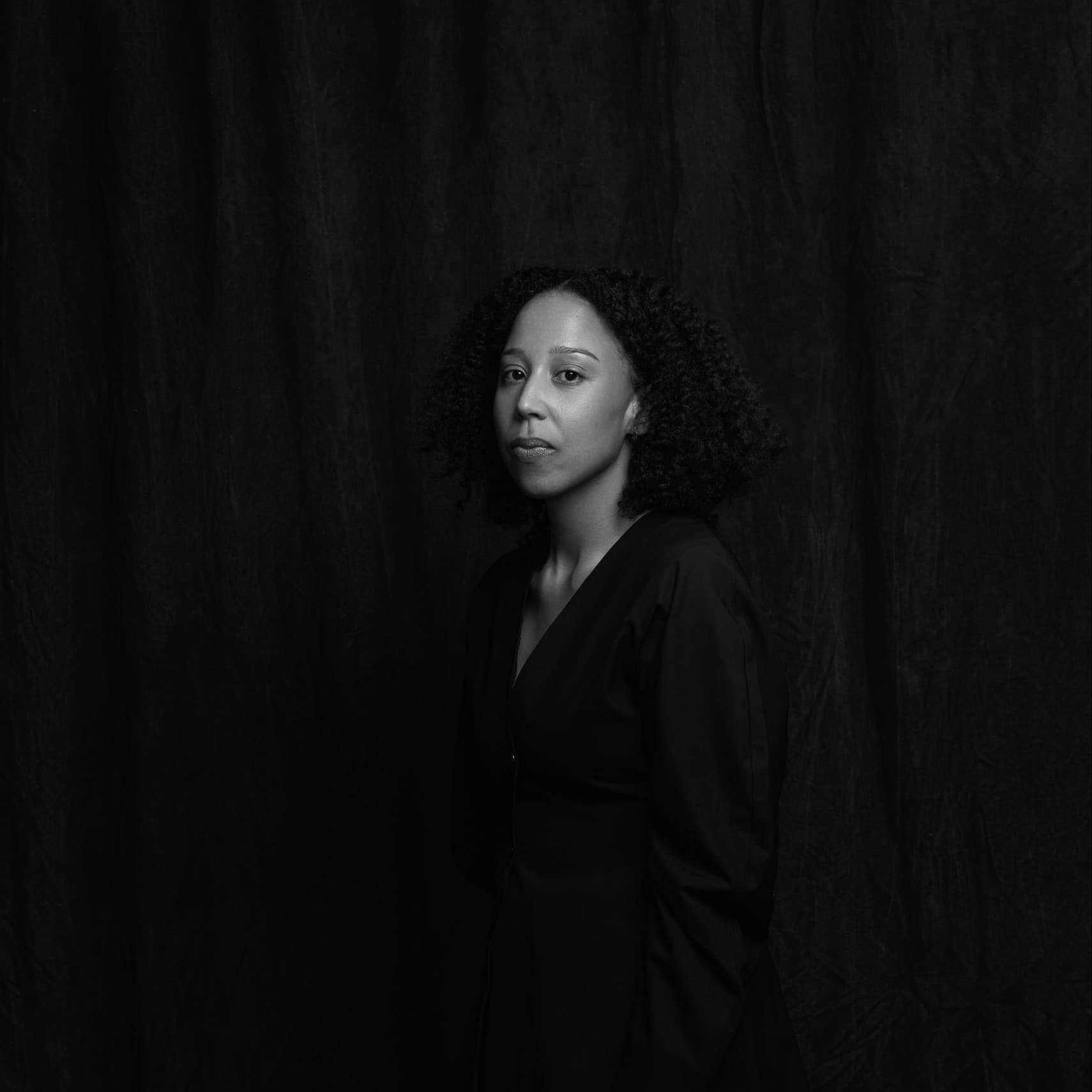 A black-and-white picture of a woman named Madeleine Hunt-Ehrlich. She is depicted standing against a dark, textured curtain background. She is facing the camera with her body slightly turned to her left. She has shoulder-length curly hair that frames her face. Her expression is calm and introspective, with a composed demeanor. She is wearing a dark, long-sleeved top barely visible against the dark background.