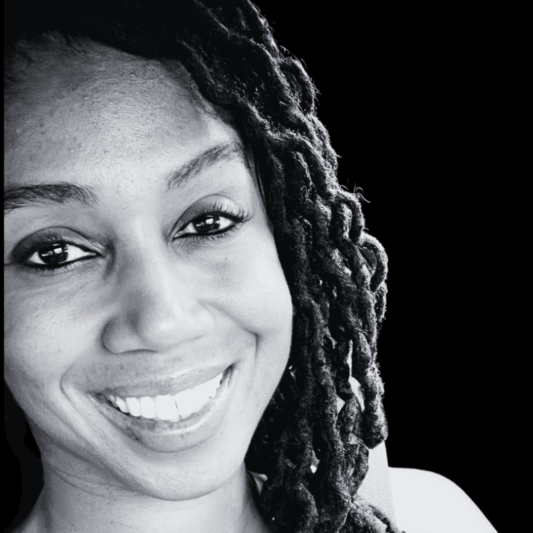 A black-and- white picture of a woman named K. Nicole Mills. She has tightly coiled, shoulder-length locks cascading down the right side of her face. She is smiling brightly, revealing straight, white teeth against a solid black background.