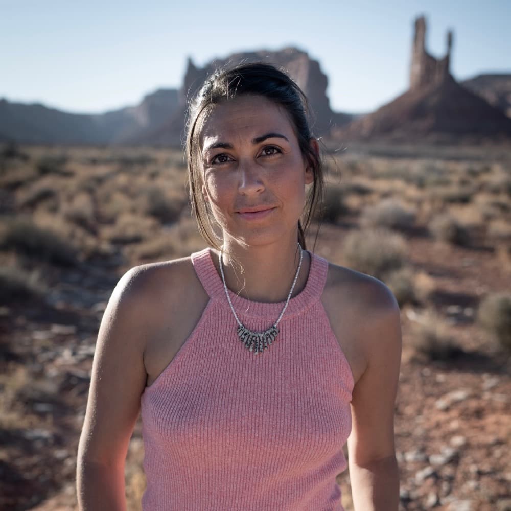A picture of a woman named Nadia Gill, standing outdoors in a desert landscape. She is centered in the frame, looking directly at the camera with a calm and neutral expression. Nadia has dark brown hair tied back in a simple ponytail. She is wearing a sleeveless, light pink knit top and a distinctive silver necklace composed of fan-like elements hanging in a semi-circle around her neck.