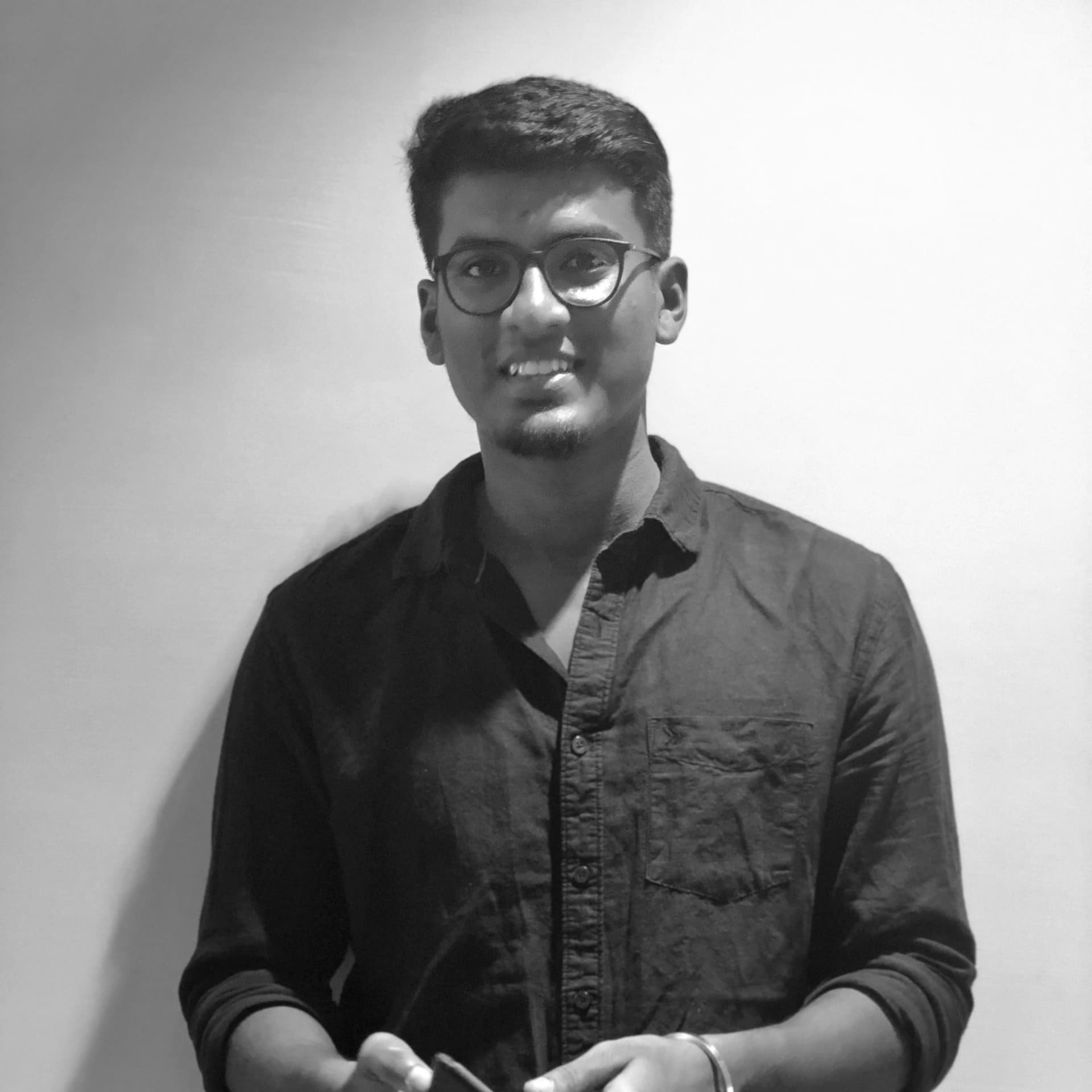 A black-and-white picture of a young man named Shiva Krish standing against a plain white background. He is captured from the waist up, wearing a dark, long-sleeved button-up shirt with the sleeves rolled up to his elbows. He has short, neatly styled hair and wears round glasses. He has a slight beard and a friendly smile with his hands positioned in front of him, holding a mobile phone.
