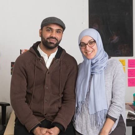 A picture of two individuals. Turab Shah, a man on the left, and Arwa Aburawe, a woman on the right, stand closely together in an office environment. Turab Shah is dressed in a brown cardigan over a white shirt and wears a flat cap. He has a neutral expression with a hint of a smile. Arwa Aburawe is wearing a light gray hijab, glasses, and a matching light gray top. She has a broad, warm smile. The background features a cluttered bulletin board filled with various colorful notes, postcards, and photos.