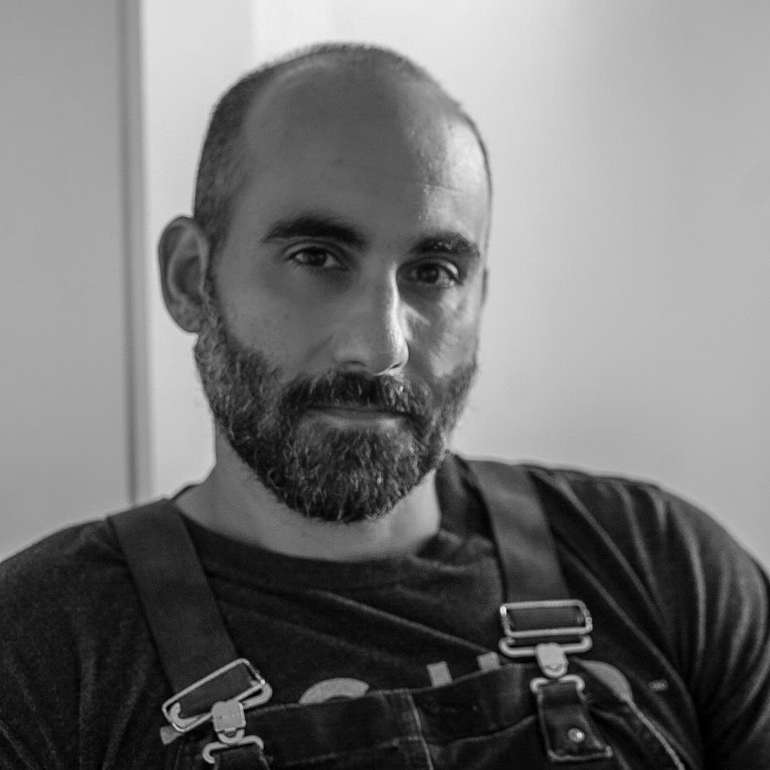 A black-and-white picture of a man named Andy Sarjahani. He is facing the camera directly and has a shaved head and a full beard peppered with gray. He is wearing dark-colored overalls over a dark shirt against a solid light background.