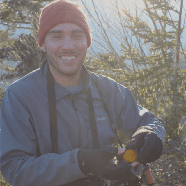 A picture of a man named Mitch Wenkus. He is in a natural setting with evergreen trees in the background, under a bright, sunny sky casting a light glow. He smiles warmly and is wearing a red beanie, a gray jacket, and black gloves. He is holding a camera that he has around his neck.