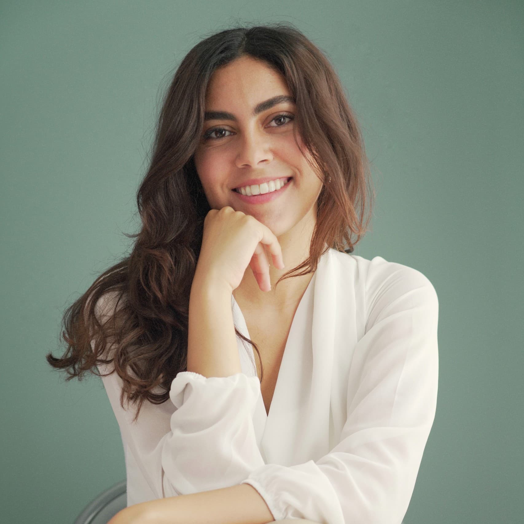 A picture of a woman named Lamees Almakkawy, seated on a chair against a muted sage green background. She is composed in a relaxed and confident pose, with her left arm resting on her knee and her right hand positioned under her chin. She is wearing a long-sleeved, flowy white blouse beige bottom. Her long, wavy brown hair cascades over her shoulders, framing her smiling face.