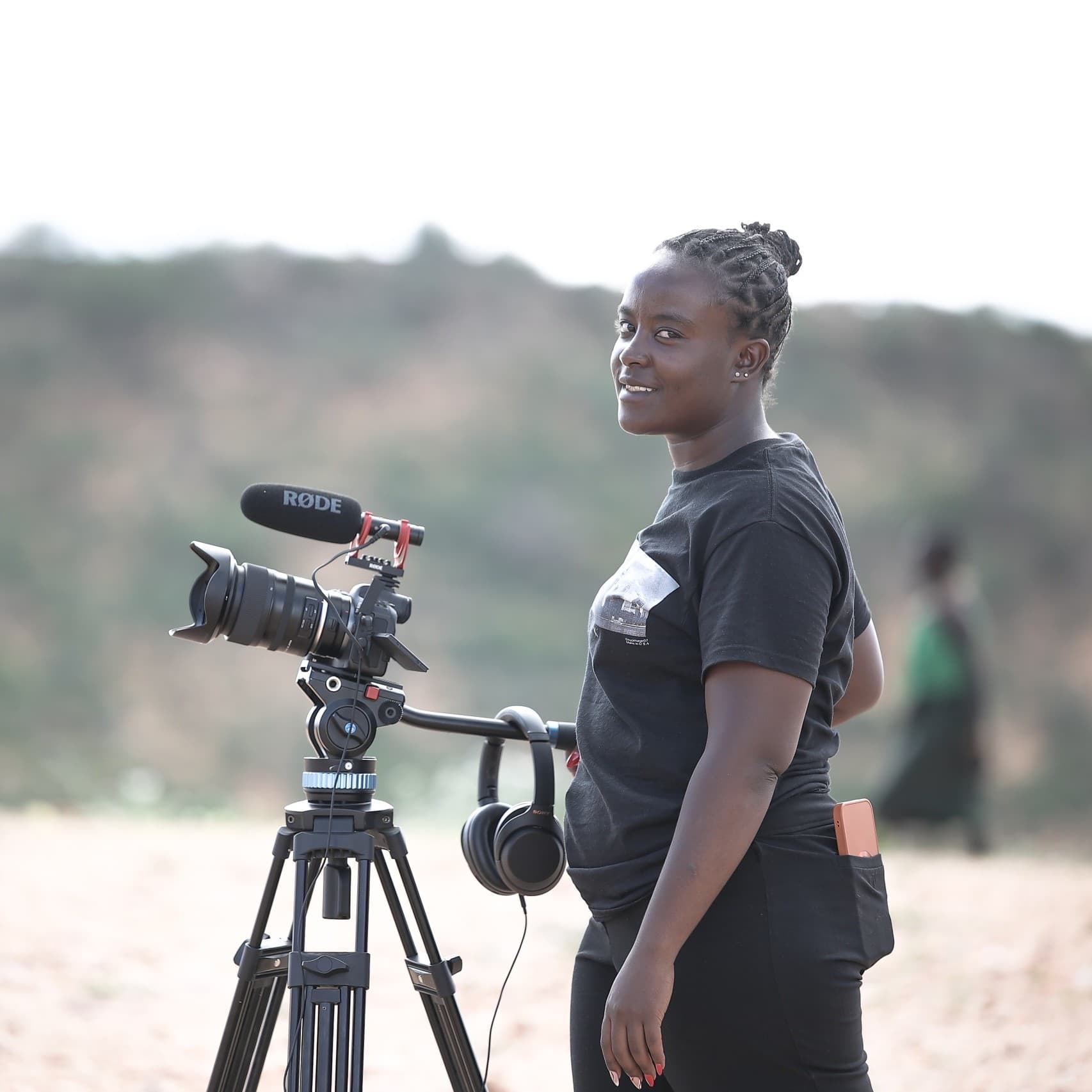 A picture of a woman named Laissa Malih, standing outdoors with a professional camera mounted on a tripod. Laissa is smiling and looking slightly over her shoulder at the camera. She has braided hair tied back and is wearing a short-sleeved black t-shirt and black pants. The background is a blurred, natural landscape with some greenery and distant hills.