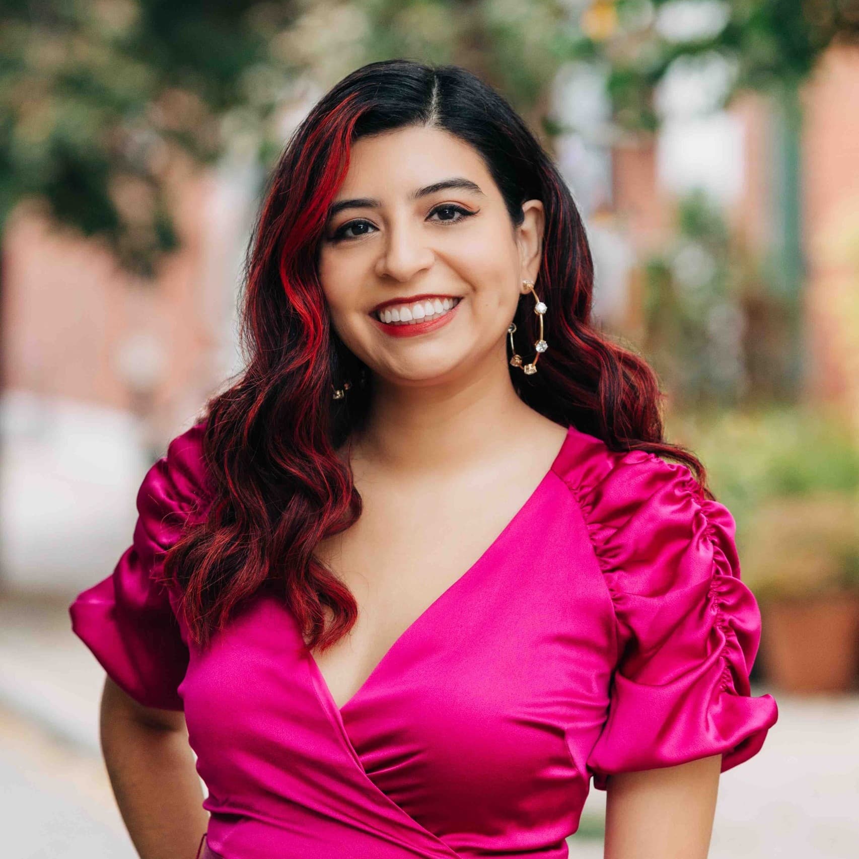 A picture of a non-binary person named Kristal Sotomayor standing outdoors. She is dressed in a vibrant fuchsia wrap dress that features puffed sleeves and a belt tied at the waist. Her dark hair, which has a red highlight on one side, falls in soft waves over her shoulders. She is smiling broadly with a friendly expression.