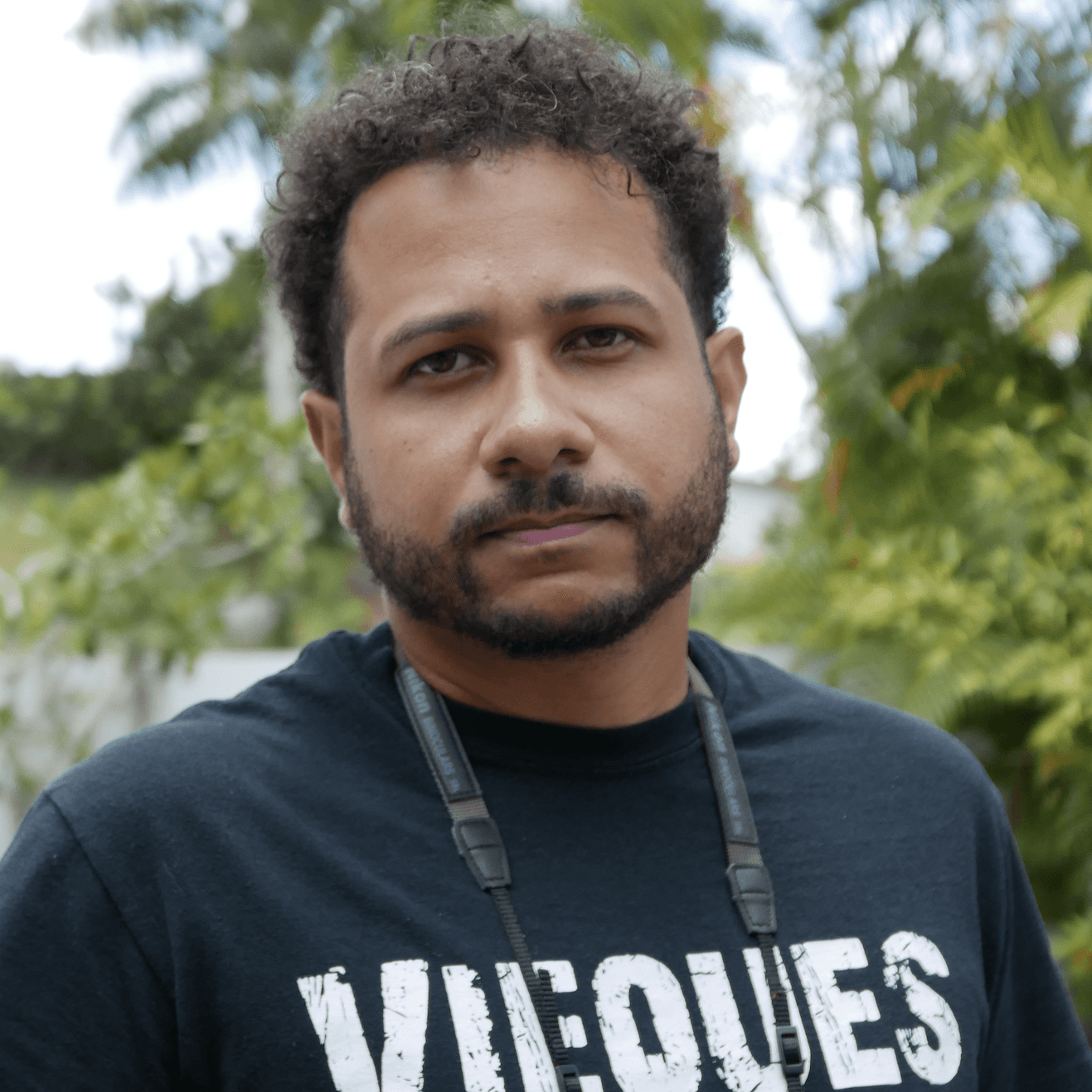 A picture of a man named Juan C. Dávila Santiago. He has short curly hair and a neatly trimmed beard. He is wearing a black T-shirt with the text "VIEQUES" printed in white across the chest. His expression is neutral, and he is against a background of lush greenery.