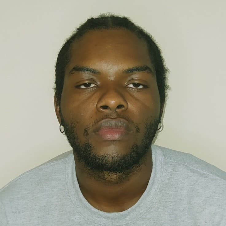 A picture of a man named Andres Mosquera. He is pictured from the chest up against a plain, light-colored background. He has a serious expression and is looking directly at the camera. Andres has a medium-length afro hairstyle, with closely cropped facial hair outlining his jaw. He is wearing a light gray sweatshirt.