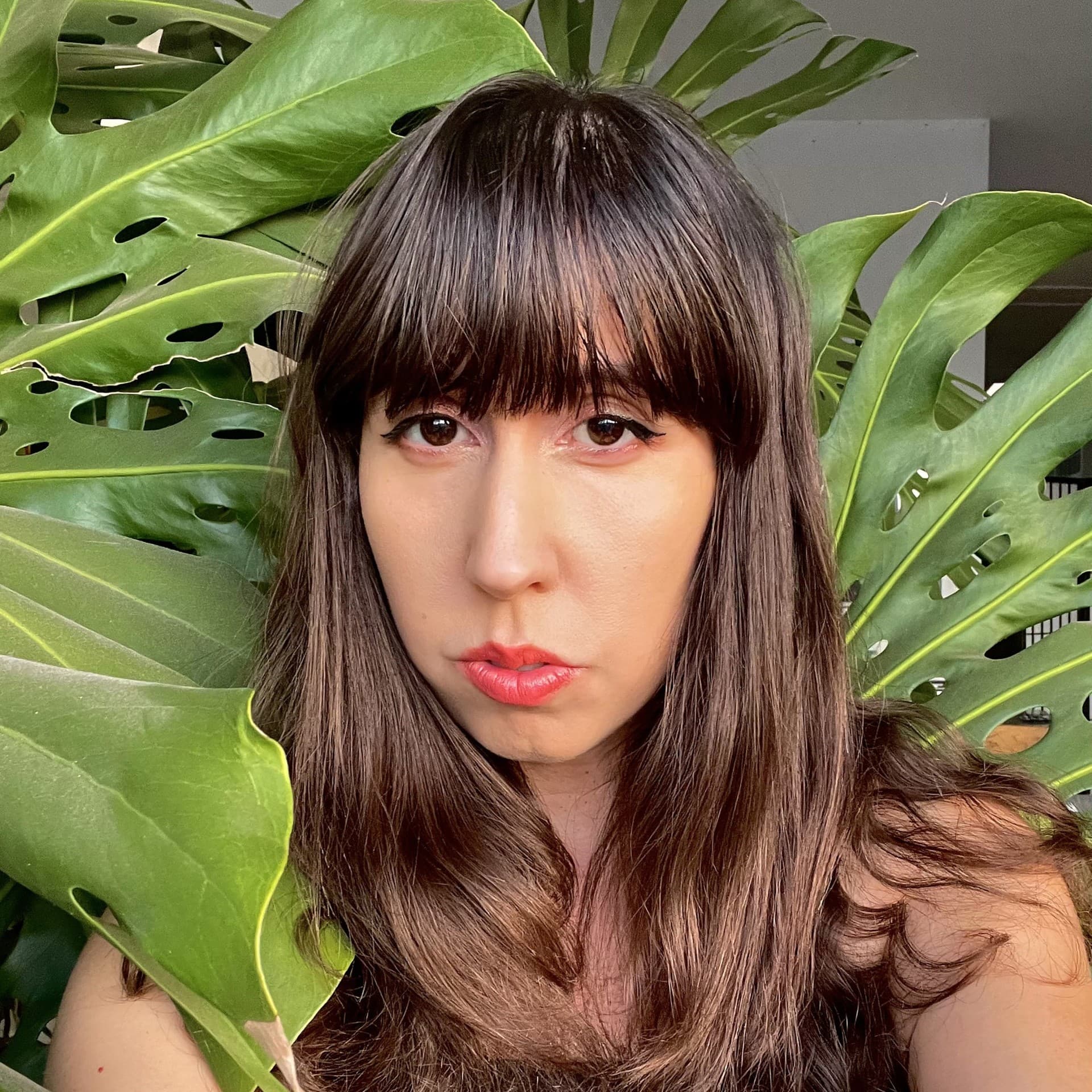 A picture of a woman named Tiare Ribeaux. She has long, slightly wavy brown hair with bangs that fall just below her eyebrows. Her expression is serious, with neutral lips accented by red lipstick, and her eyes feature dark eyeliner. She is surrounded by large, vibrant green Monstera leaves with distinctive holes and perforations.