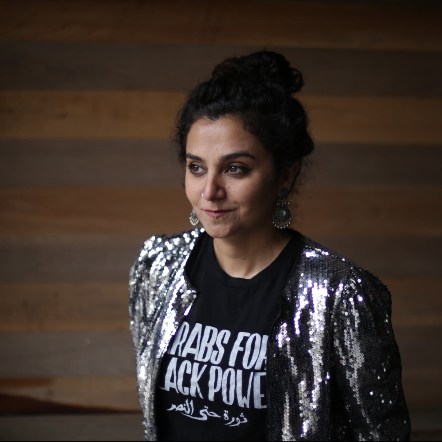 A picture of a woman named Alia Haju. She is standing indoors against a background of wooden planks with alternating shades of brown. Alia has dark, curly hair pulled up into a bun and wears large, intricate silver earrings. She is dressed in a shiny, silver sequin jacket over a black T-shirt that reads "ARABS FOR BLACK POWER" in bold white letters, along with a line of Arabic text beneath.