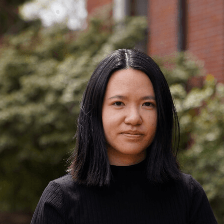 A picture of a woman named Bea Hesselbart. She is standing outdoors with a softly blurred background of greenery and a red brick building. Bea has shoulder-length straight black hair and is wearing a black top that blends seamlessly with her dark hair. Her expression is calm and composed, with a faint, gentle smile.