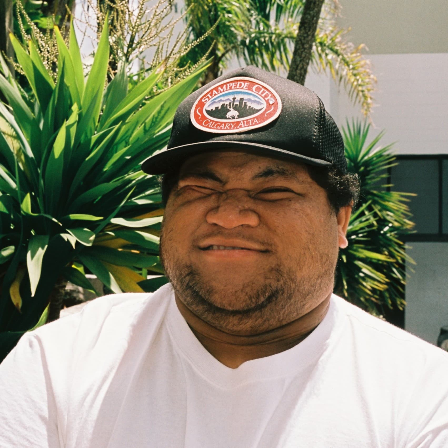 A picture of a man named Laman Time. He is outdoors and is positioned in front of a background filled with large, green, spiky leaves of tropical plants. He is wearing a white T-shirt and a black baseball cap with an emblem that reads "Stampede City Calgary, Alta." The man has a neutral expression, with his eyes squinting.