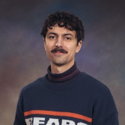 A picture of a man named Charles Frank. He has curly dark hair with a mustache and is centered against a backdrop of a smooth gradient transitioning from dark brown to purple hues. He has a calm and slightly smiling expression. He is wearing a dark blue sweater with the word "BEARS" in bold letters, highlighted by orange and white stripes. Underneath the sweater, he has a black and pink striped turtleneck.