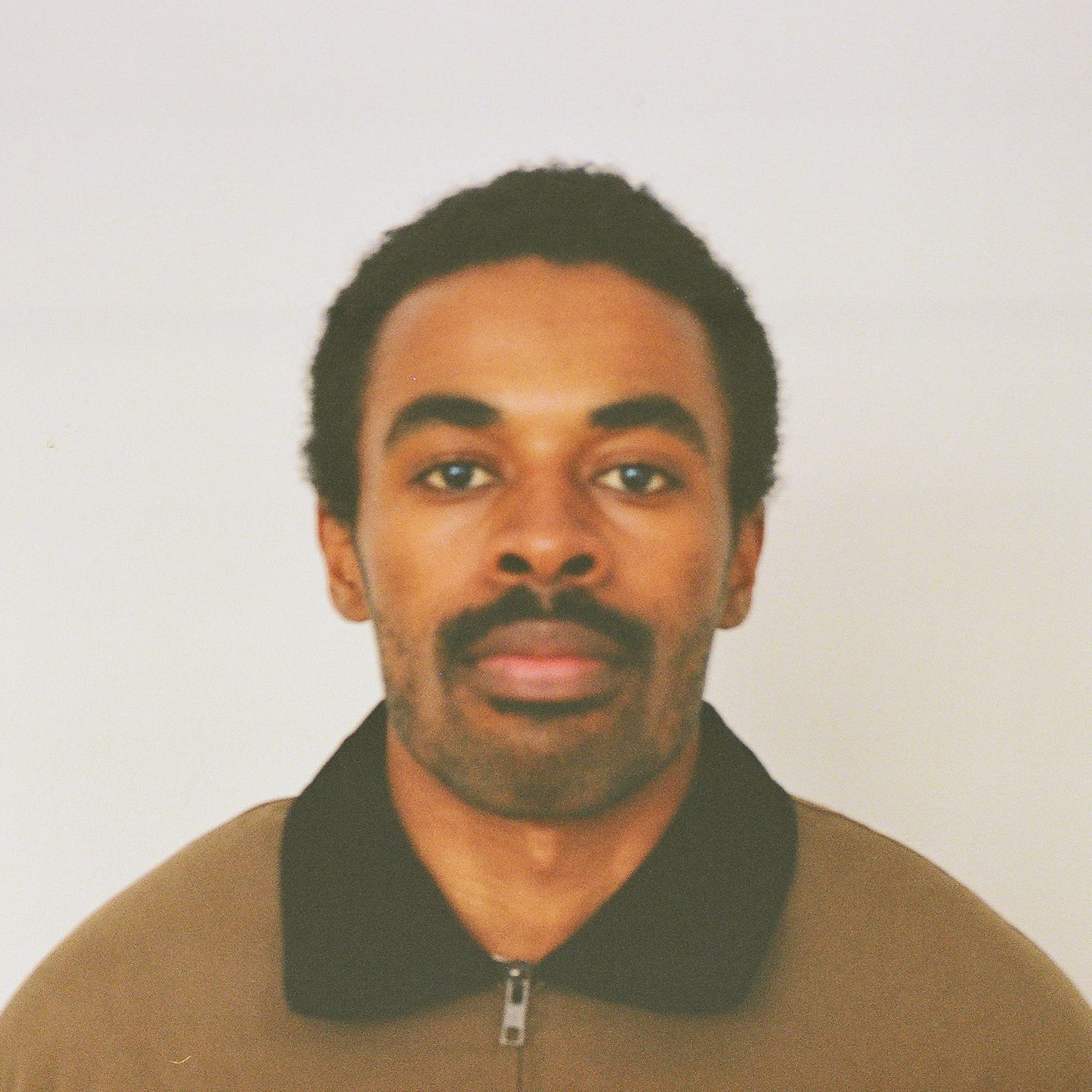 A picture of a man named Blair Barnes. He is positioned against a plain, light-colored background and has short, curly hair and a neatly trimmed mustache. He is wearing a brown zip-up jacket with a black collar. His expression is neutral, with a calm and composed demeanor.