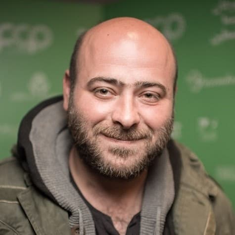 A picture of a man named Nidal Badarny. He has a bald head and a neatly trimmed beard with a friendly, subtle smile that suggests warmth. He is wearing a dark gray jacket layered under a green coat, both of which look slightly worn and casual, suitable for cool weather. The blurred background is a green backdrop with logos and text.