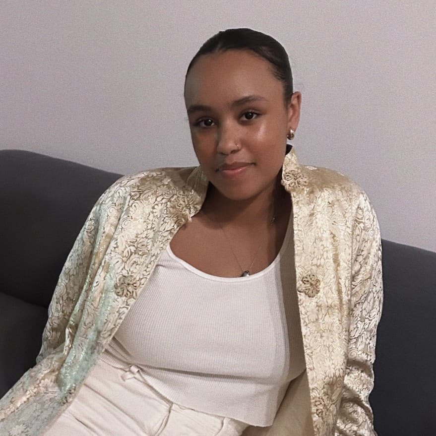 A picture of a woman named Mahsati Fidel Moorhead. She is seated on a dark gray sofa against a pale, neutral background. Mahsati is casually leaning back with one elbow resting on the arm of the sofa. She has a calm, relaxed expression on her face with a faint smile. Her hair is neatly tied back, and she is wearing hoop earrings. She is dressed in a white, sleeveless top and high-waisted white pants. Over her outfit, she wears a luxurious, cream-colored, long jacket with intricate, floral embroidery.