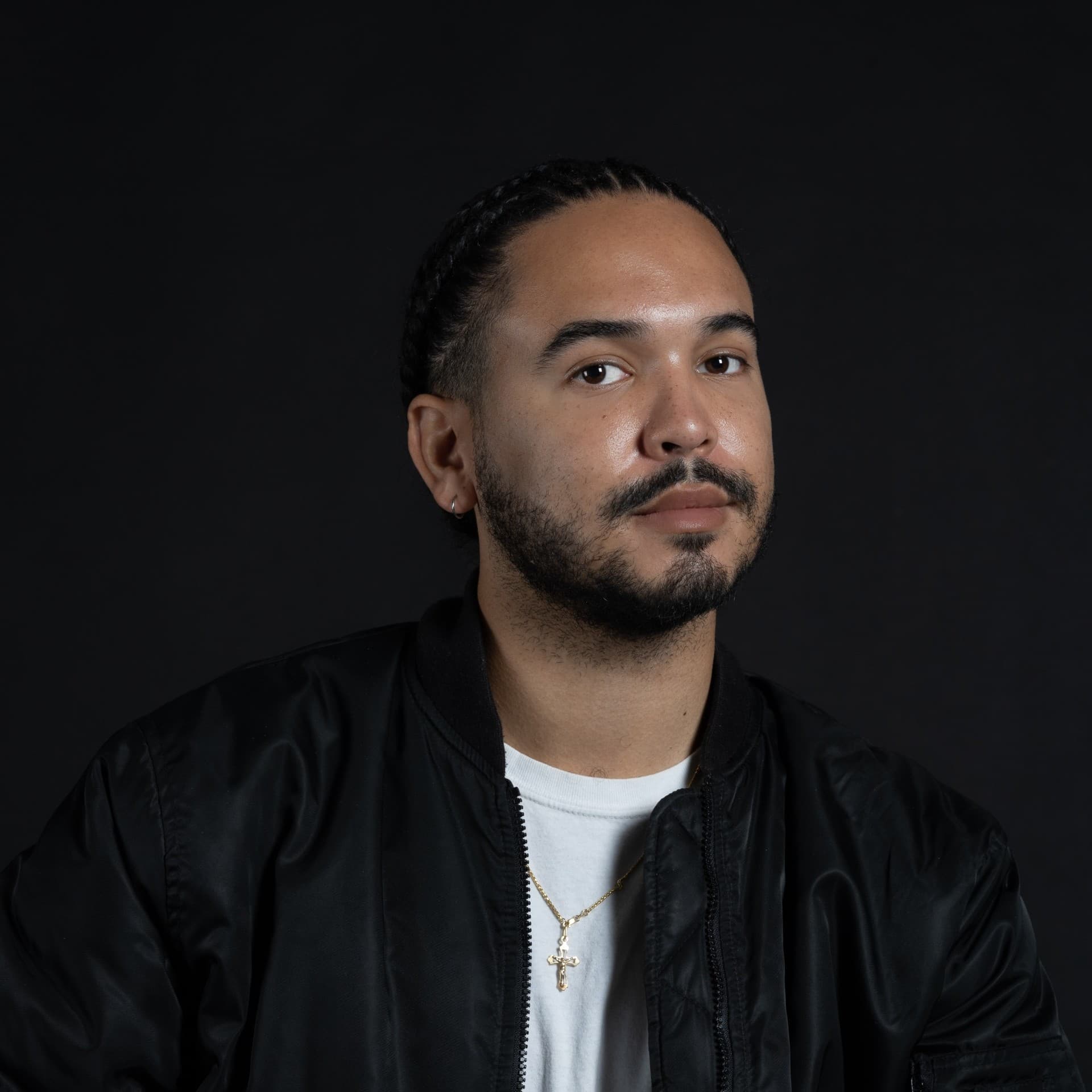A picture of a man named Joseph Douglas Elmhirst against a dark background. He has a composed expression with a subtle smile on his face, eyes looking slightly to the side. His hair is styled in neatly braided cornrows. Joseph is wearing a black bomber jacket over a white t-shirt. Around his neck, he has a delicate gold chain with a small cross pendant.