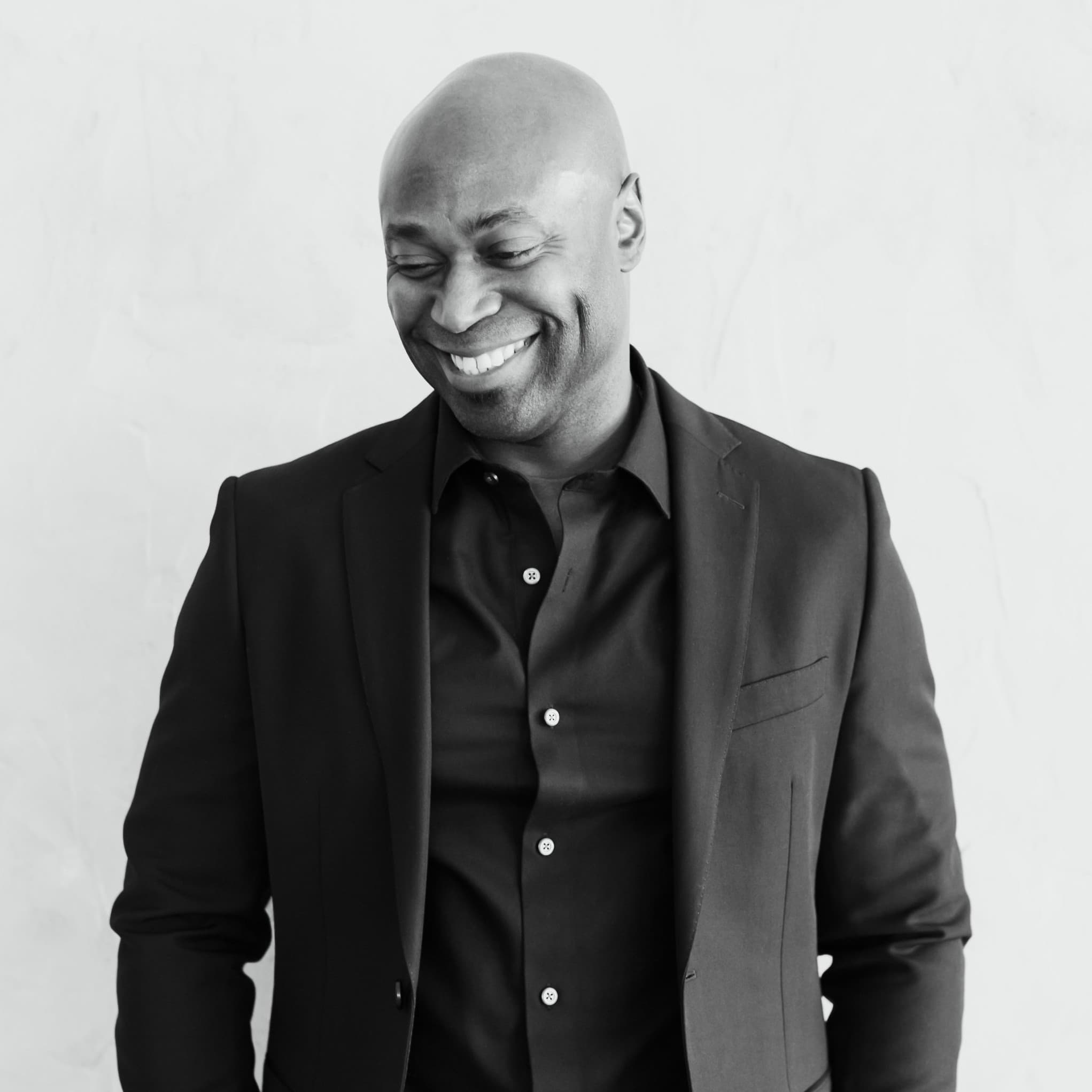 A black-and-white picture of a man named Anslem Richardson against a plain light-colored wall. He is smiling warmly while looking downwards. He is dressed in a dark, tailored suit with a matching button-up shirt underneath. The suit jacket has a simple, sleek design with a notched lapel and the shirt is neatly buttoned up to the collar. The man is standing with his hands casually resting in his pockets. His head is bald, and his facial expression exudes a serene and content demeanor.